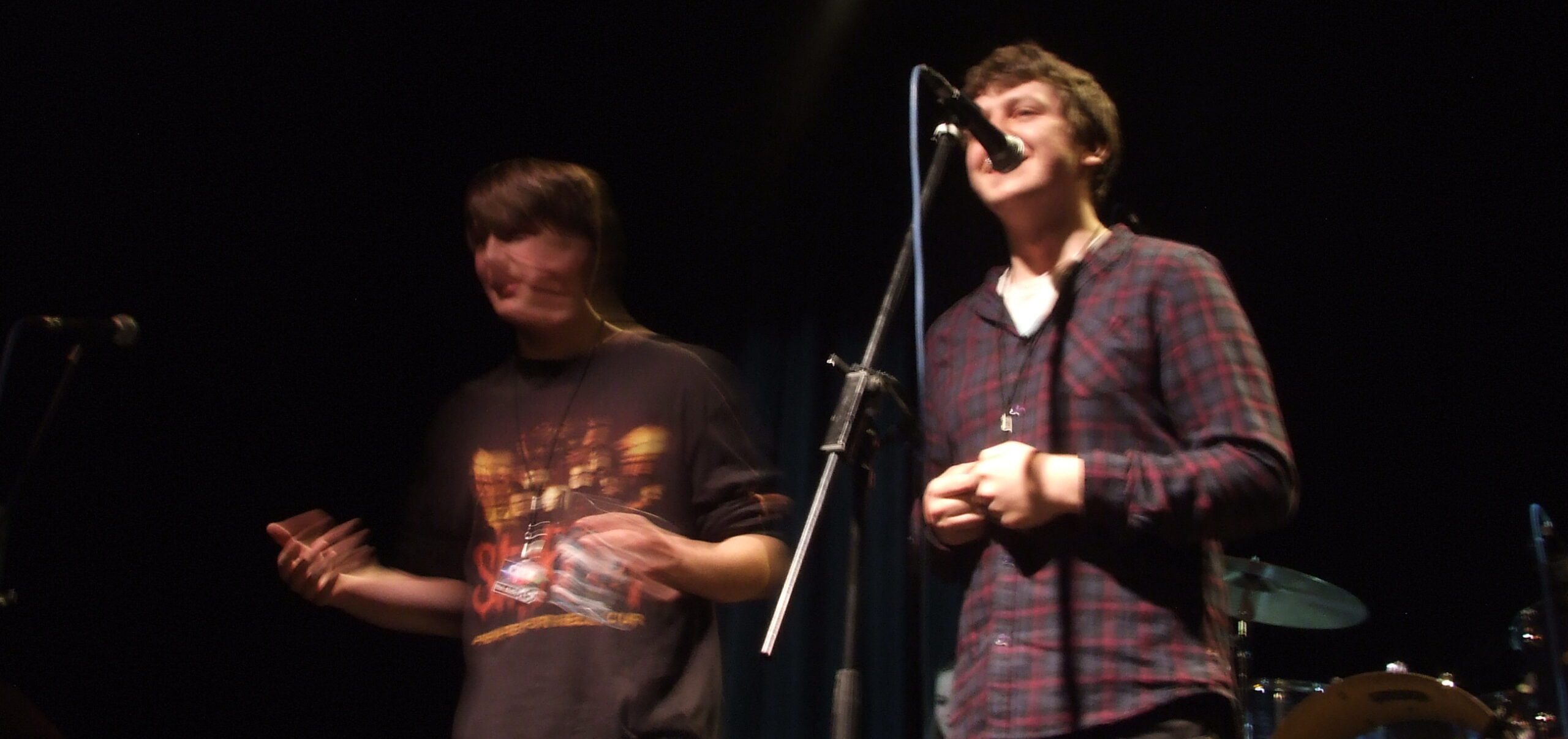 A teenage boy stands under a spotlight on a dark stage, singing into a microphone in front of him. Next to him, another teenage boy is slightly blurred as he dances.